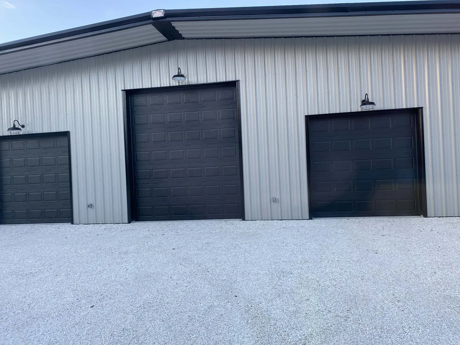 Three black short-panel overhead doors in a ribbed steel shop wall, gooseneck barn lights over each opening and a fresh gravel apron