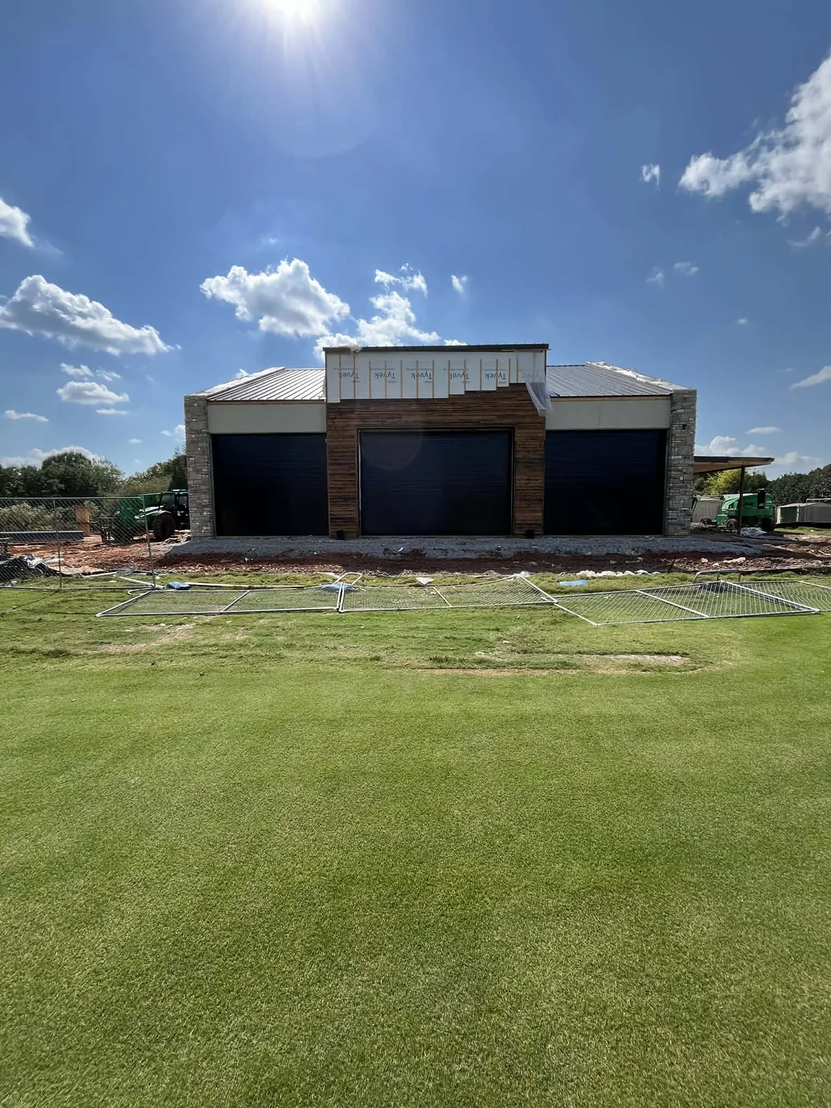 Three black rolling steel doors hung in a stone and timber equipment building, the roof trim still wrapped and turf running to the slab