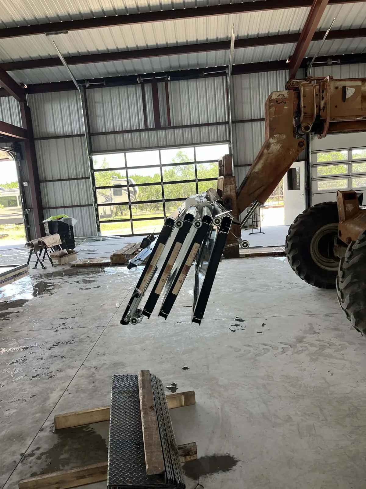 Track, brackets and door sections staged on the slab ahead of a hang, a telehandler parked alongside and a full-view door already set in the far opening