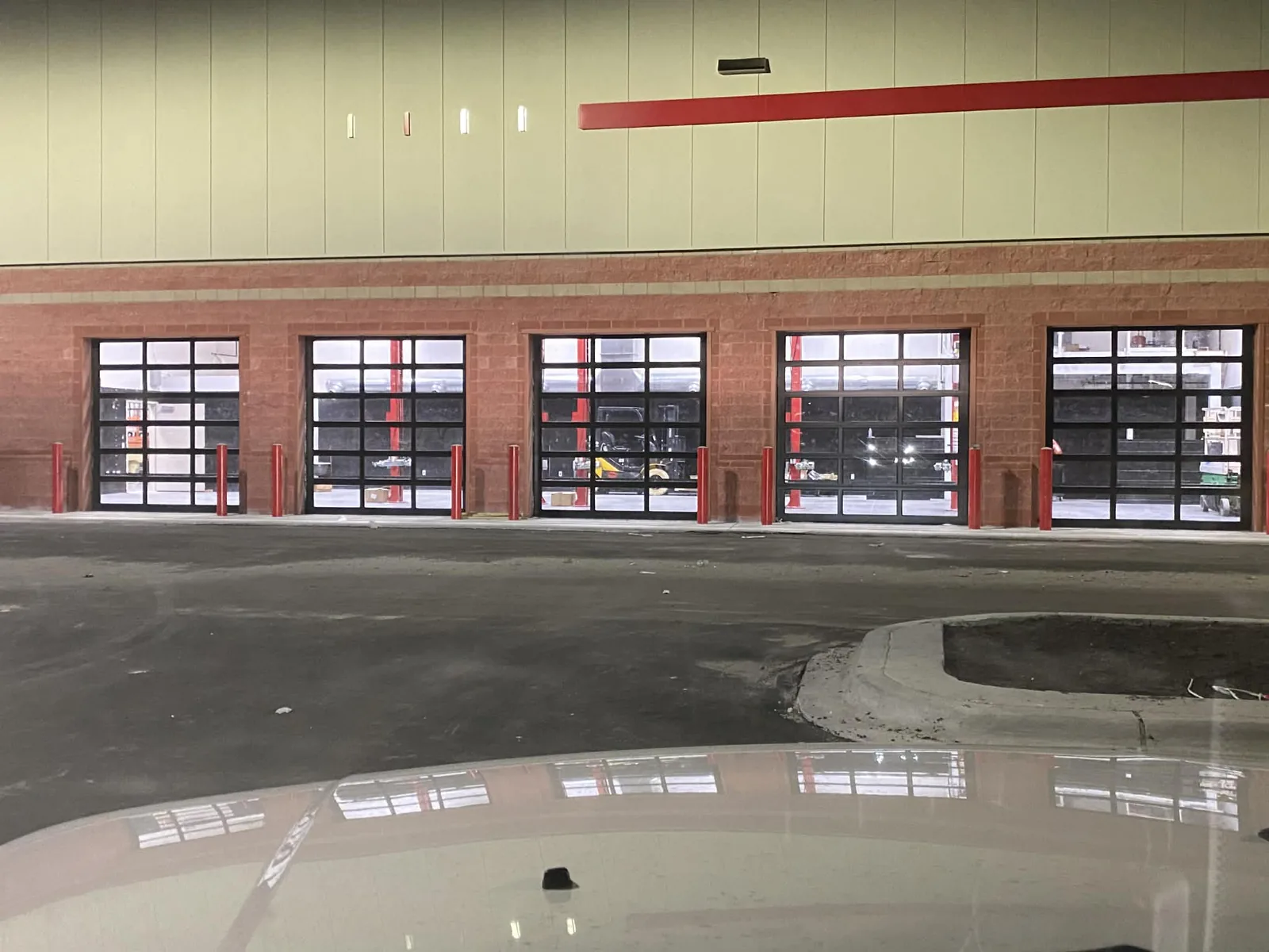 Five full-view aluminum and glass sectional doors closed across a brick service building after dark, red bollards guarding each jamb