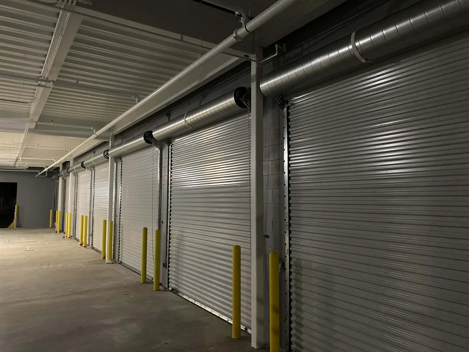 A long corridor of galvanized rolling steel doors with pull chains at every opening, yellow bollards along the slab and spiral duct running the length of the bay