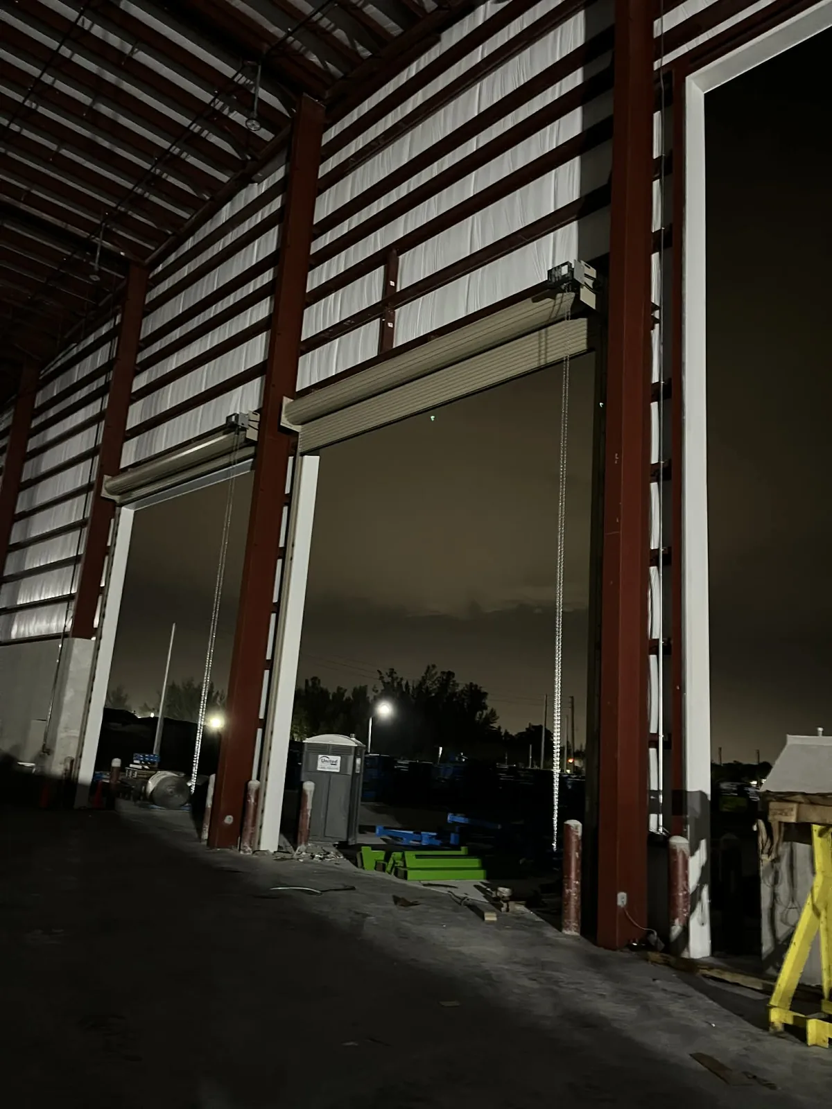 Two rolling steel doors set high in a red-frame steel building and left open overnight, hand chains hanging clear of the guides