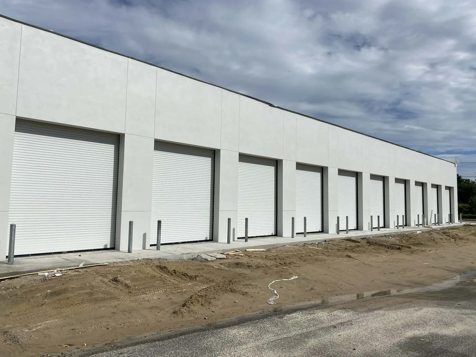 Ten white roll-up doors set in a tilt-wall warehouse elevation, bollards in front of each opening and the apron not yet paved