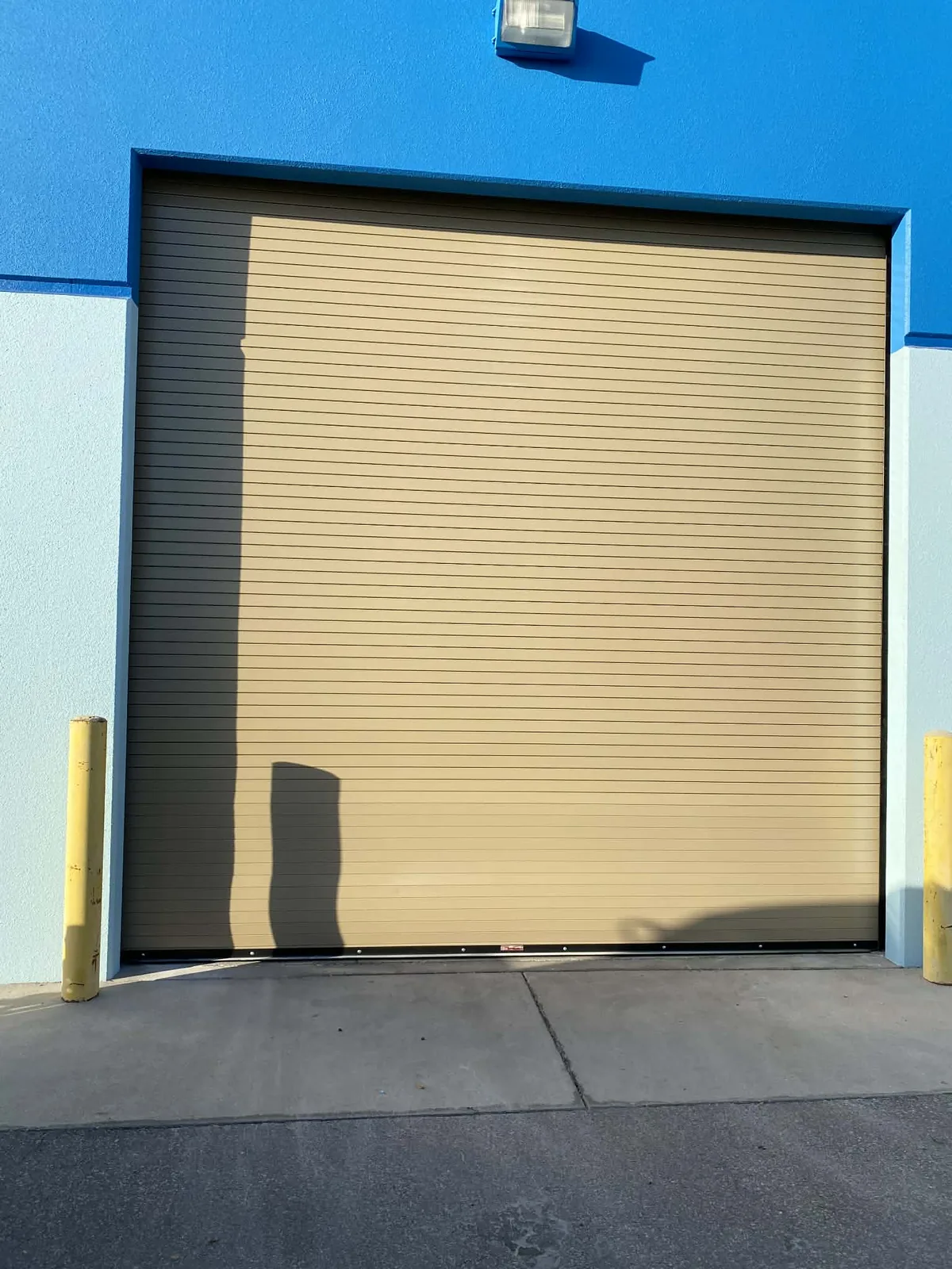 Almond rolling steel service door in a masonry commercial building, bollards set either side of the opening and a wall pack above the hood