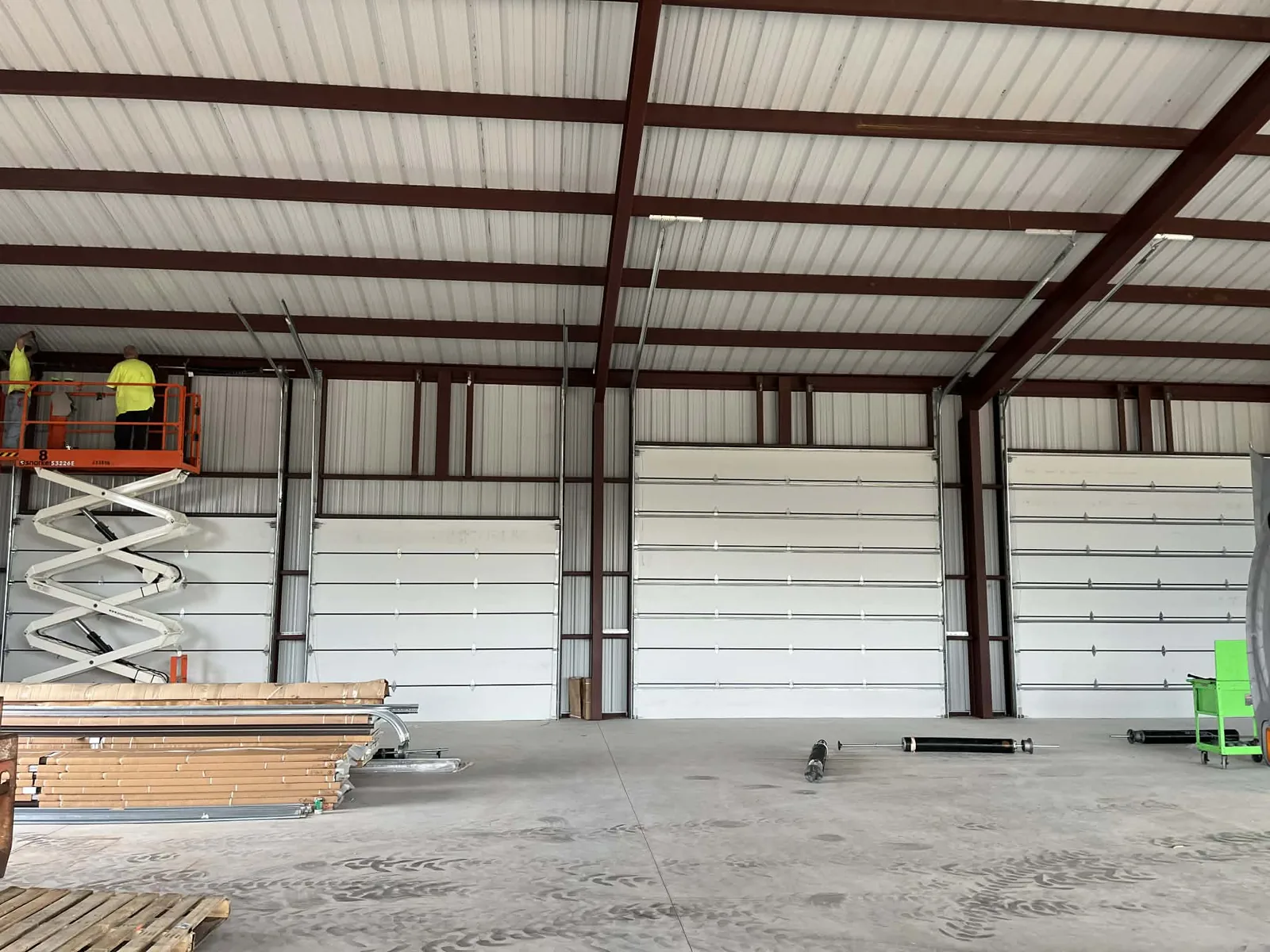 Three sectional doors hung inside a red-steel building while two technicians work the header from a lift, torsion tubes and track still laid out on the slab