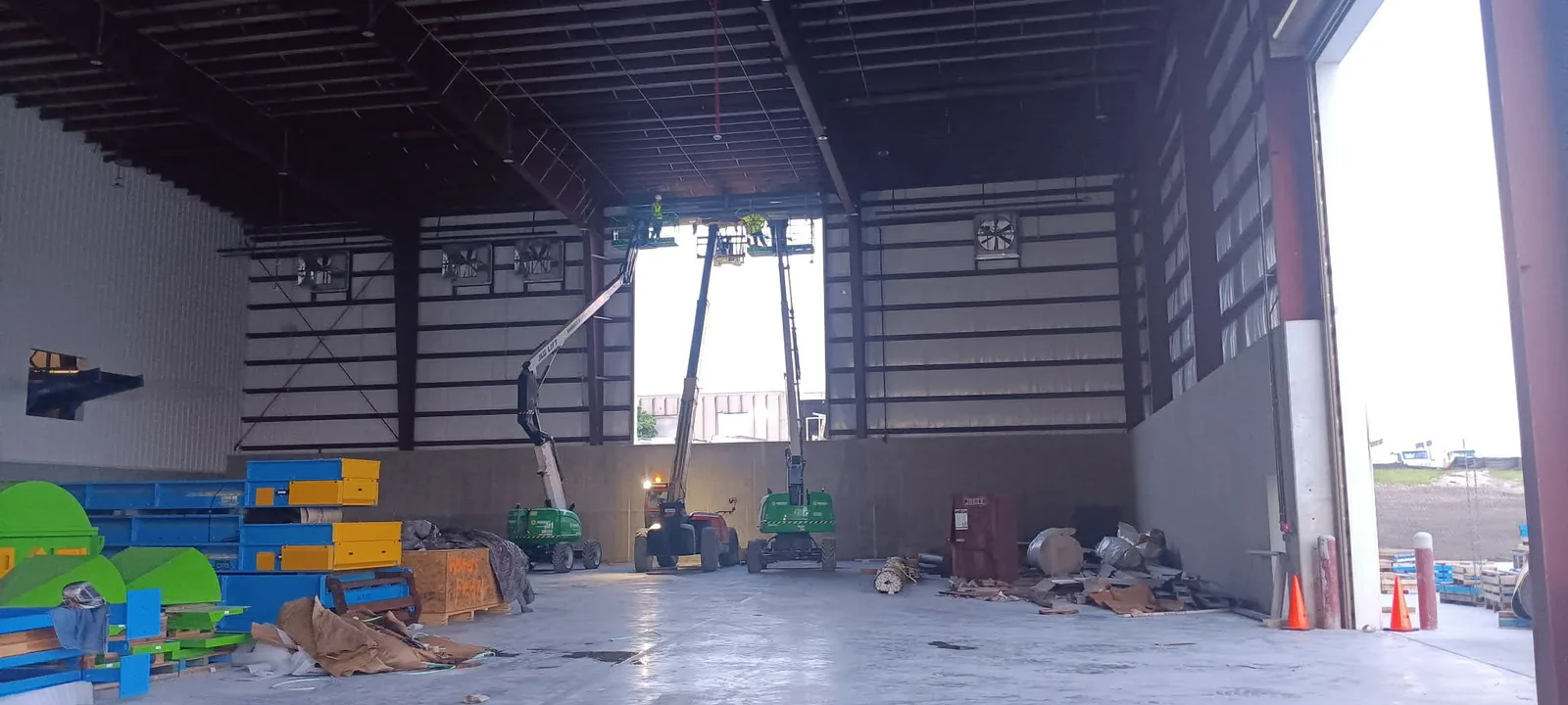 High-bay warehouse shell with the drive-through opening framed out, boom lifts staged under the roof deck and exhaust fans set in the end wall